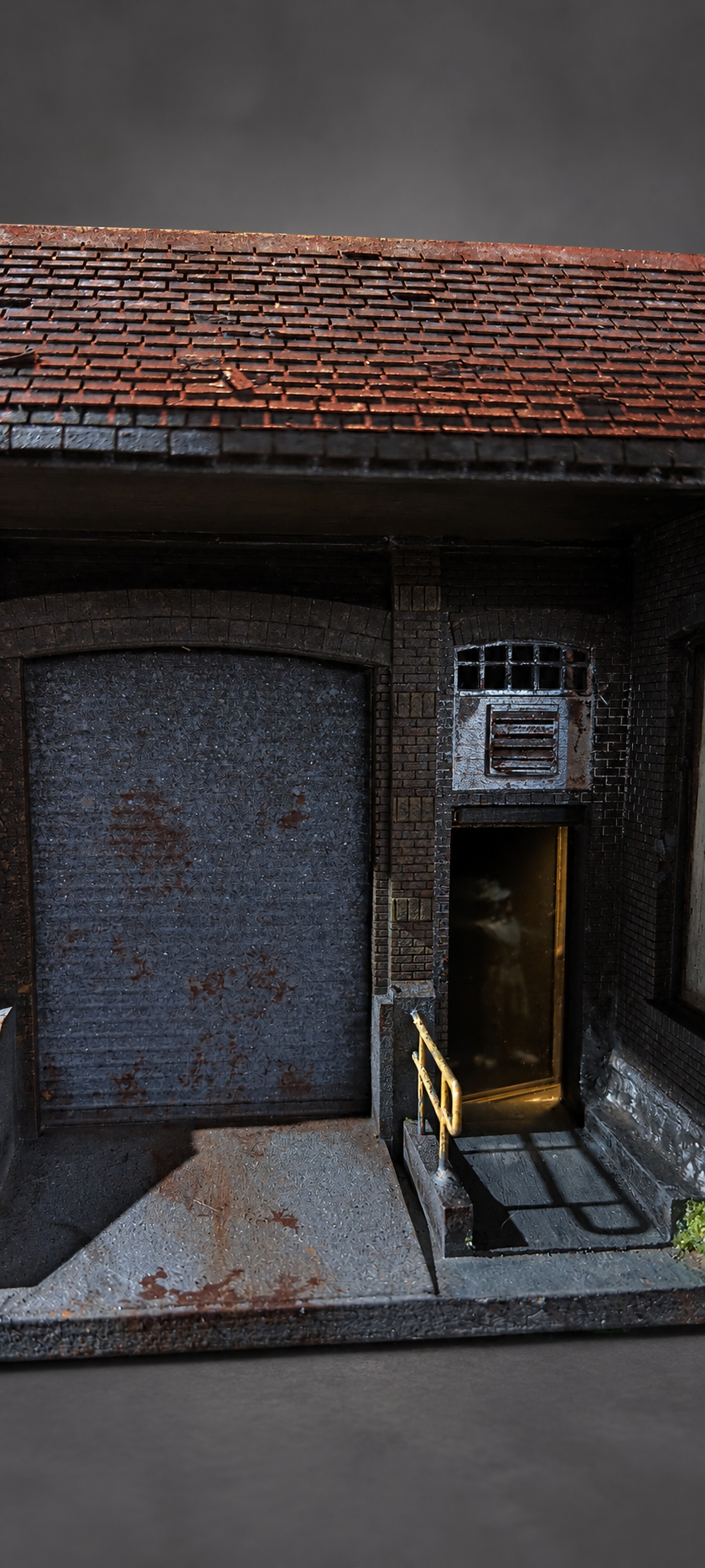 A miniature model of a weathered brick building with a rusted metal garage door, a small open doorway with a yellow railing, and a tiled roof under a dark gray sky.