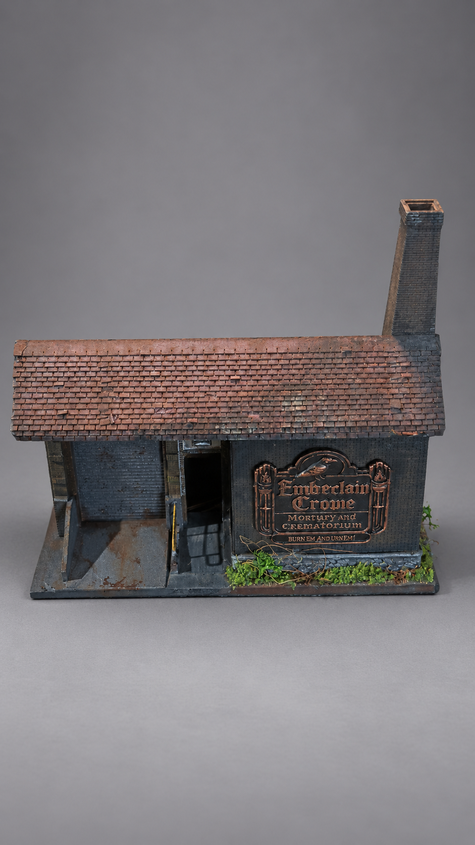 A miniature model of a brick warehouse building with a tall chimney, dark gray walls, a brown tiled roof, and a decorative sign on the front, photographed on a neutral gray background.